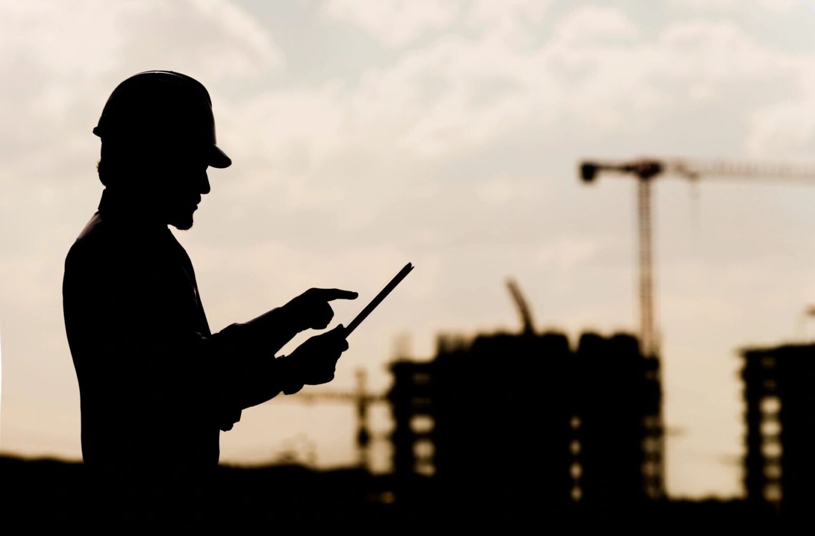 Silhouette of a construction worker with a hard hat using a tablet at a construction site during sunset.