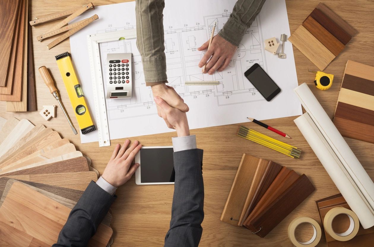 Two professionals shaking hands over a table scattered with architectural plans and construction materials.
