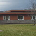 Modern single-story building with a mix of wood and stone facade, featuring a flat roof and large windows, set against an overcast sky.