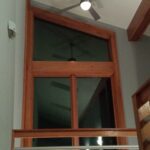 A high interior wall with a large triangular window, a ceiling fan above, and a wooden railing in the foreground.
